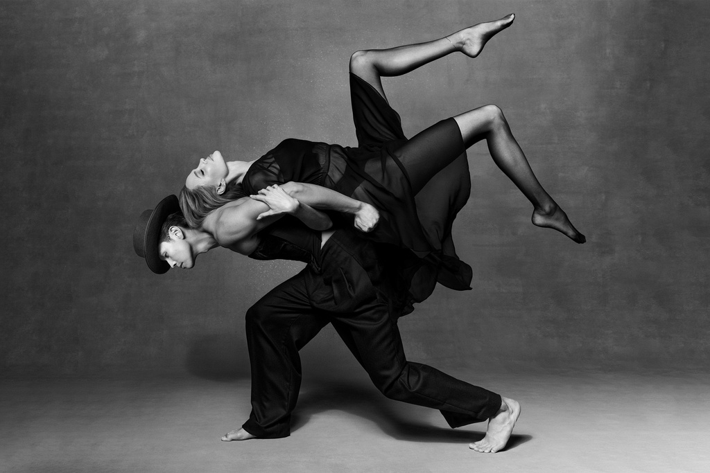 Two dancers in black clothing strike a dynamic pose in a studio, one supporting the other in a grayscale studio shot.