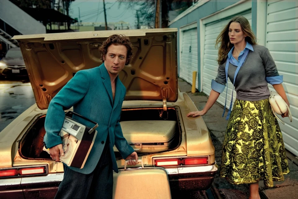Retro-styled fashion shot featuring a man in a teal jacket and woman in a floral skirt by a gold car with suitcases, evoking a vintage vibe.