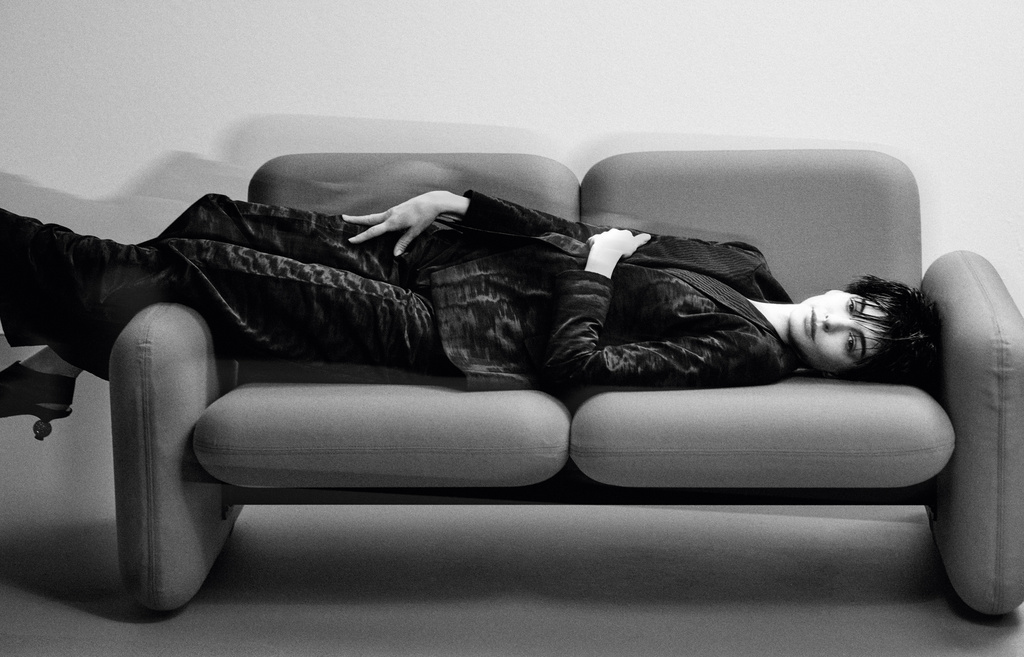 Woman lying on a grey couch wearing a black suit. The black and white image creates a high contrast, classic and elegant aesthetic.
