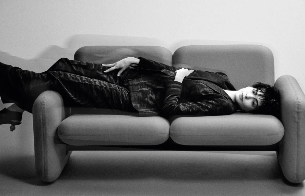 Woman lying on a grey couch wearing a black suit. The black and white image creates a high contrast, classic and elegant aesthetic.