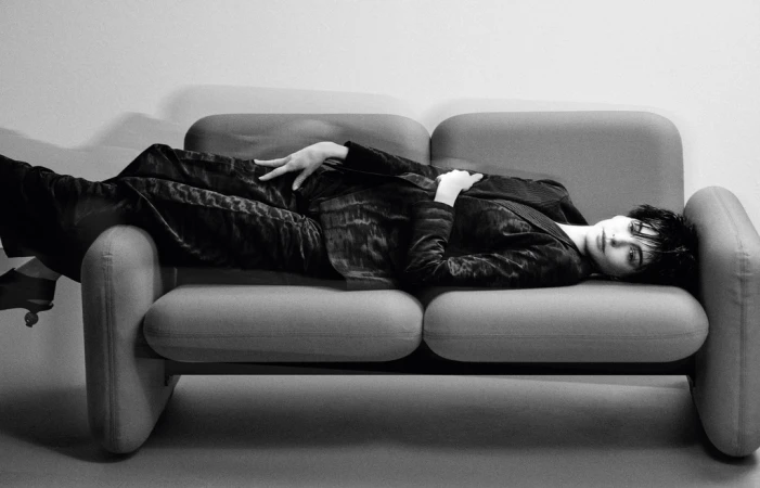 Woman lying on a grey couch wearing a black suit. The black and white image creates a high contrast, classic and elegant aesthetic.