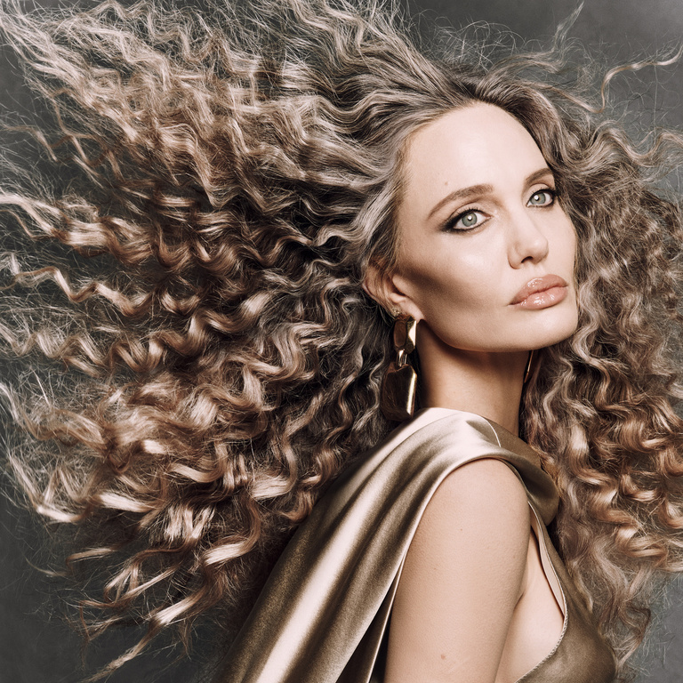 Close-up of a woman with big curly hair, gold dress and gold statement earrings against a grey background, beauty photography.