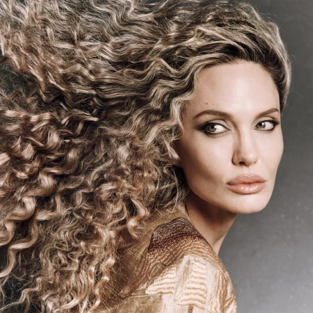 Close-up portrait of a woman with voluminous flowing curly blonde hair and dramatic eye makeup against a textured dark background