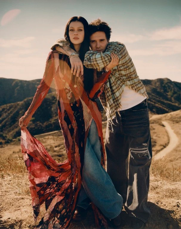 Two people posing outdoors on a dry hillside, a woman in a long patterned sheer dress and a man in a plaid shirt.