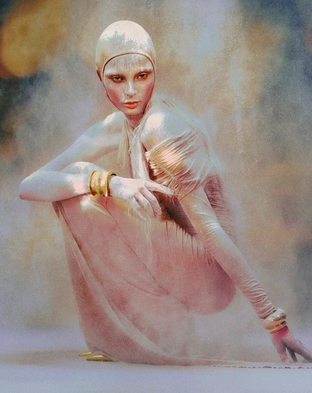 Model in metallic gold cap and body paint with avant-garde makeup crouching in an ethereal dusty setting.