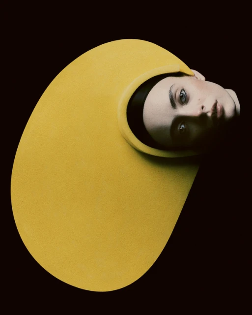 Portrait of a person wearing a yellow helmet-like garment against a black background, featuring editorial styling and studio photography.