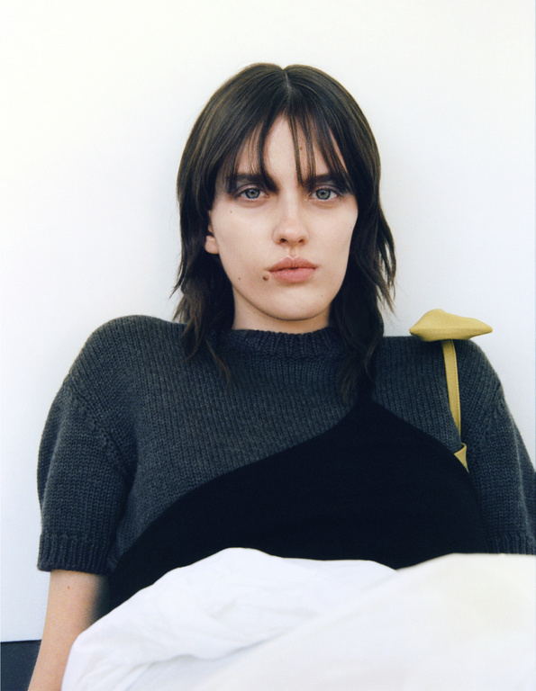 Close-up portrait of a young adult woman with shoulder-length brown hair, wearing a sweater and black top, against a white background.