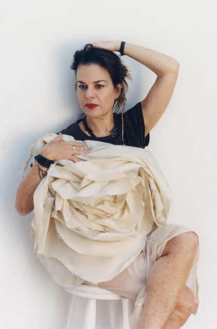 Adult female sitting on a white stool, holding a large, light-colored flower-like fabric sculpture in front of a white background.
