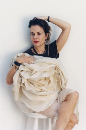 Adult female sitting on a white stool, holding a large, light-colored flower-like fabric sculpture in front of a white background.