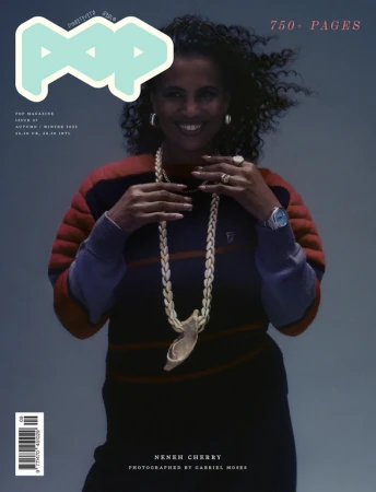 Portrait of Neneh Cherry wearing a striped sweater and shell necklace against a grey studio backdrop