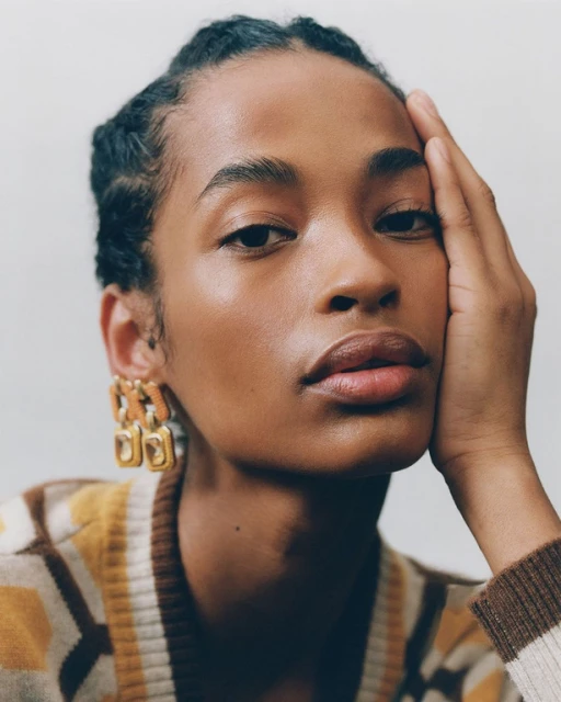 Close-up portrait of an adult woman with hand on face, wearing statement earrings and a patterned cardigan against a neutral background.