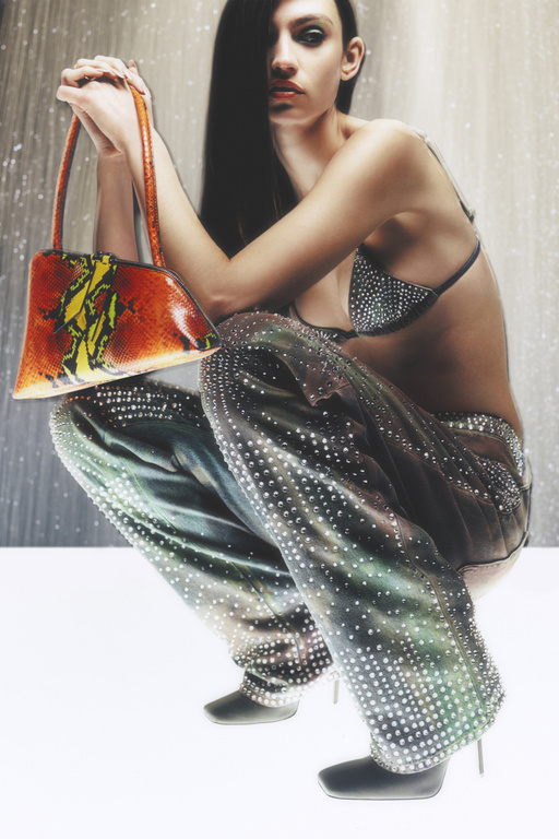 A woman in embellished pants and top poses crouching, holding an orange patterned bag, against a muted backdrop, fashion style.