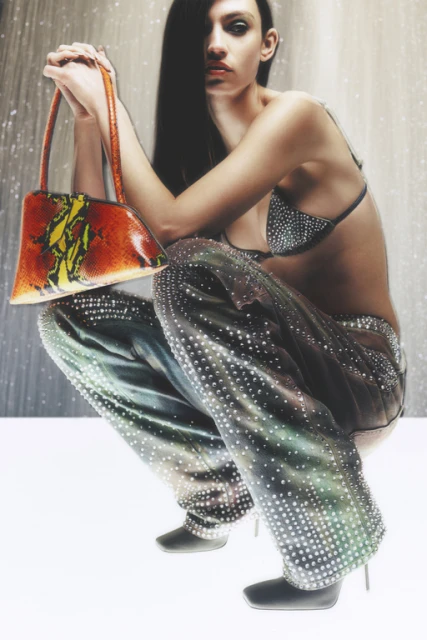 A woman in embellished pants and top poses crouching, holding an orange patterned bag, against a muted backdrop, fashion style.