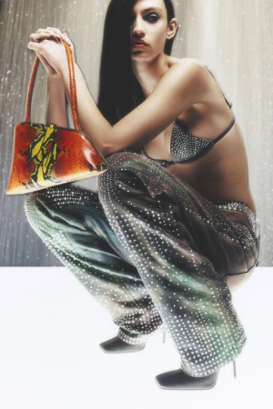 A woman in embellished pants and top poses crouching, holding an orange patterned bag, against a muted backdrop, fashion style.