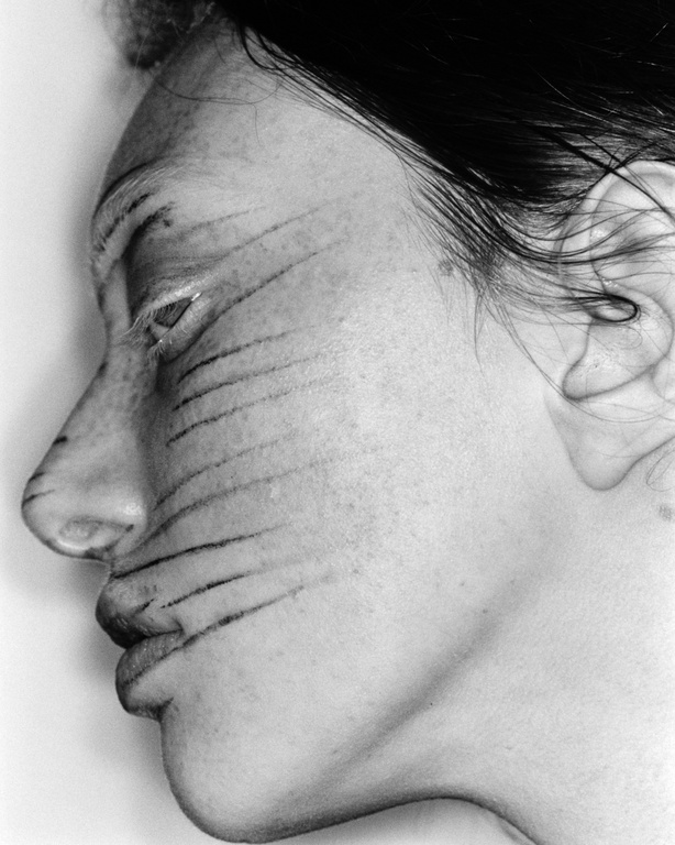 Monochrome portrait of a woman's face in profile, featuring striking black stripes painted across her skin, creating an artistic, graphic effect.