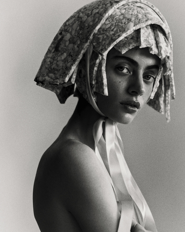 Monochrome portrait of a young woman with light skin wearing a textured headdress with ribbons, shot in a photo studio.