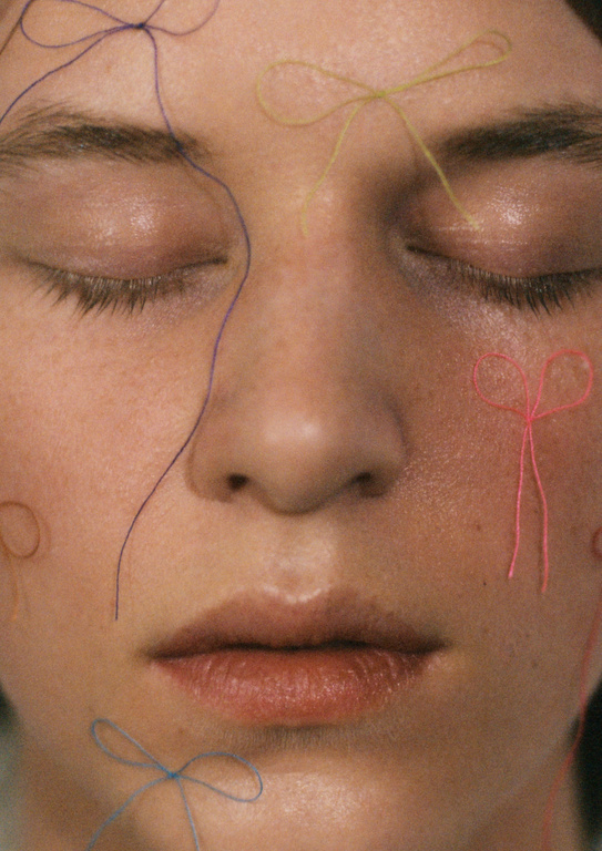Close-up portrait of a woman with closed eyes and fair skin decorated with colorful thread shaped into bows, creating a whimsical aesthetic.