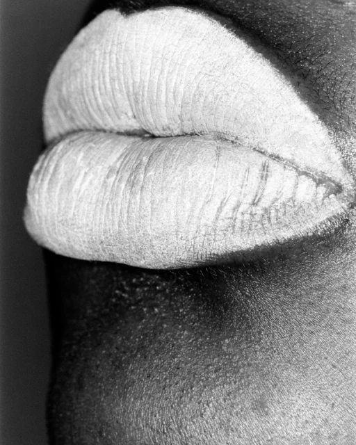 Close-up of a woman's mouth featuring lips with white makeup, set against a dark complexion in a black and white studio photograph.