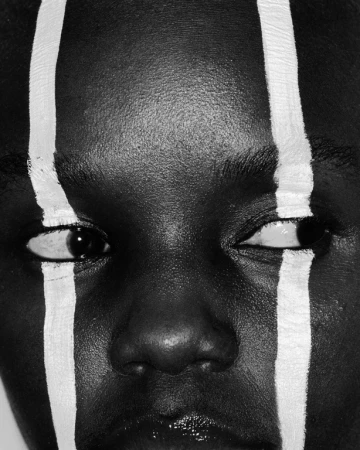 Close-up portrait of a Black person's face with bold vertical white paint stripes running across forehead and eyes by make-up artist Erin Green.