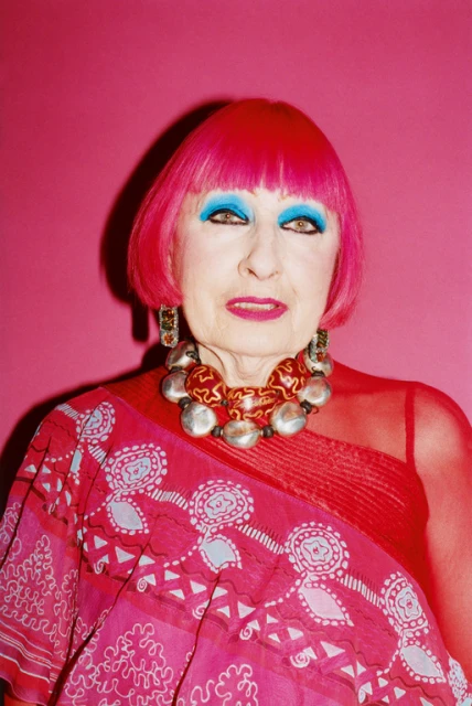 Portrait of an older adult woman with bright pink hair, blue eyeshadow, and a patterned pink top against a vibrant pink backdrop.