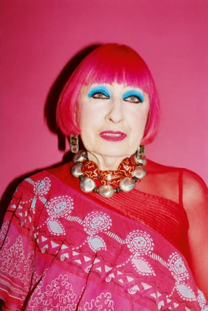 Portrait of an older adult woman with bright pink hair, blue eyeshadow, and a patterned pink top against a vibrant pink backdrop.