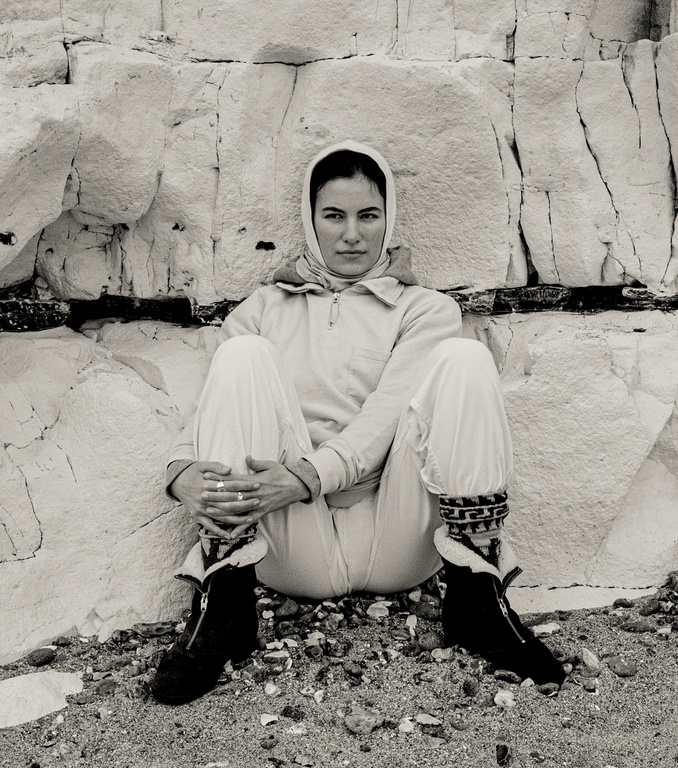 Black and white portrait of a woman wearing a headscarf and a jumpsuit sitting in front of a light-colored stone wall.