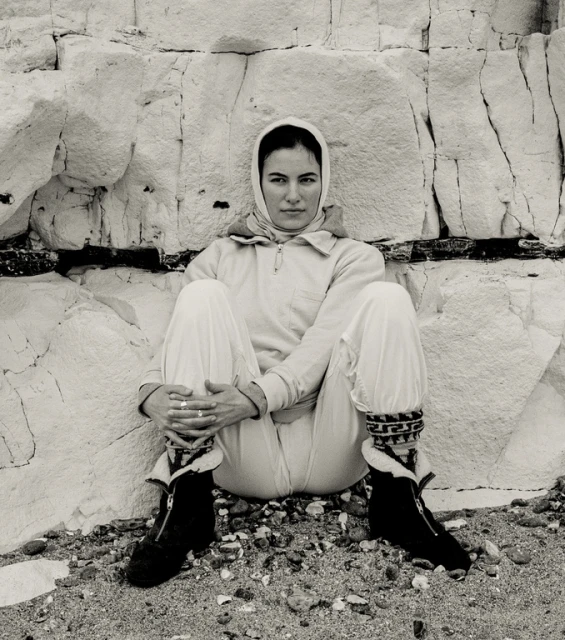 Black and white portrait of a woman wearing a headscarf and a jumpsuit sitting in front of a light-colored stone wall.