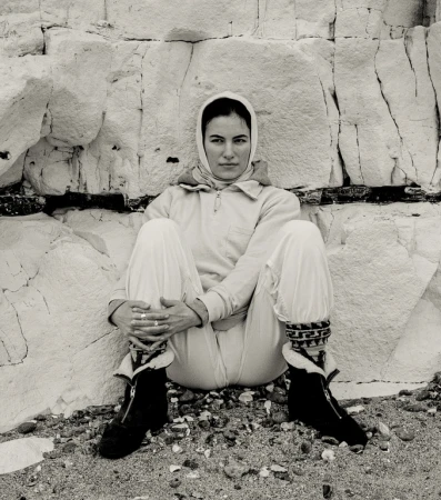 Black and white portrait of a woman wearing a headscarf and a jumpsuit sitting in front of a light-colored stone wall.
