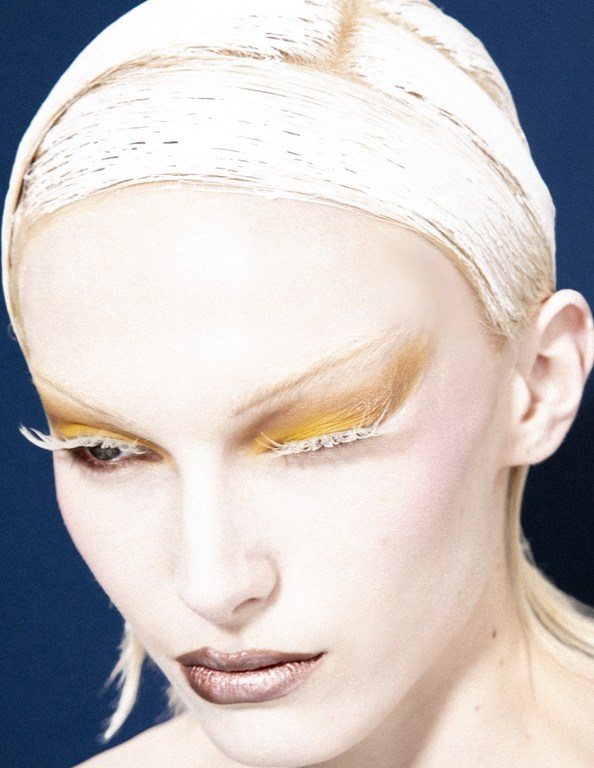 Close-up studio shot of a woman with bleached blonde hair and dramatic eye makeup featuring yellow eyeshadow and white lashes.