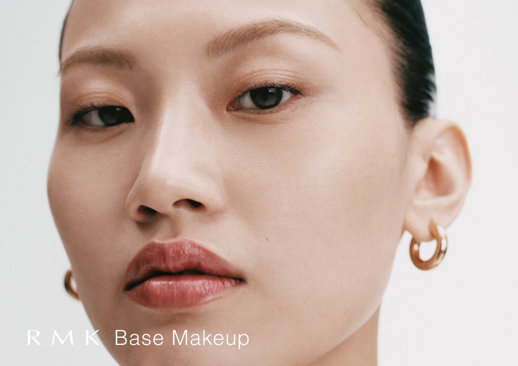 Close-up portrait of an Asian woman with radiant skin, minimal makeup, gold hoop earrings, set against a clean white backdrop.