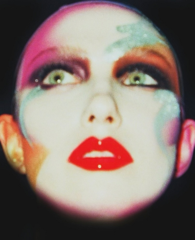 Close-up beauty portrait of a model with vibrant colorful makeup and bold red lips against a black background.