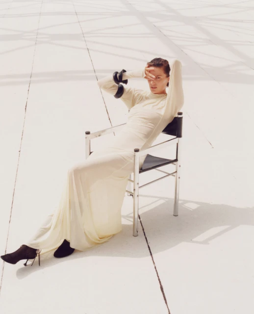 Adult woman in a long dress sits in a chair outdoors, shielding her eyes against a white background with linear details.