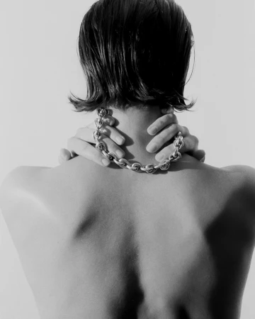 Close-up back view of a person wearing a chunky silver chain necklace with hands touching the neck, set against a plain background.