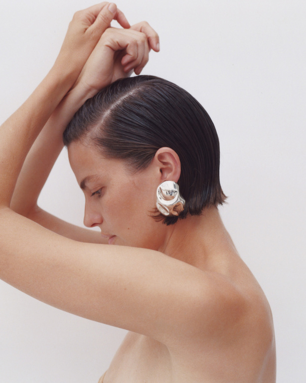 Profile shot of a woman with short dark hair and silver earrings, posing with her hands above her head against a white backdrop.