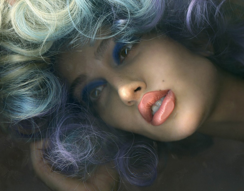 Close-up portrait of a woman with blue eye makeup and light and blue tinted curly hair, lying down, with a soft diffused lighting.