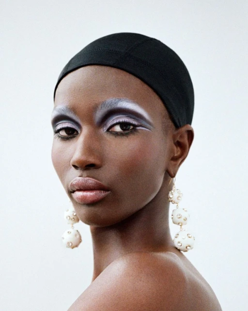 Close-up studio shot of a Black adult woman with dramatic silver eye makeup, wearing a black head covering and large dangling earrings.