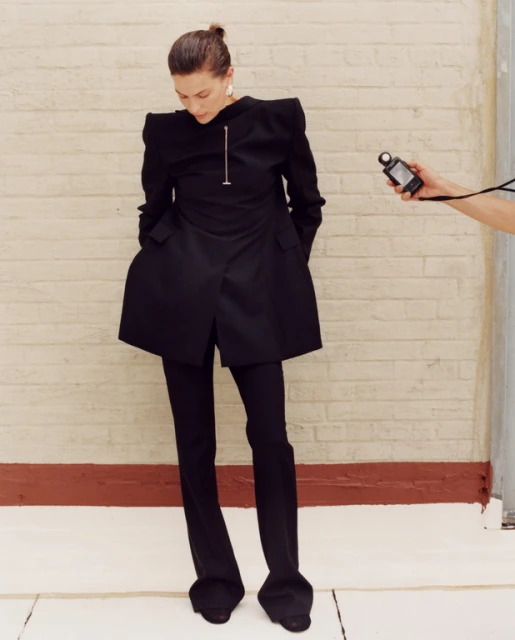 Stylish woman in a black pantsuit strikes a pose against a brick wall, with a light meter held by a person out of frame.