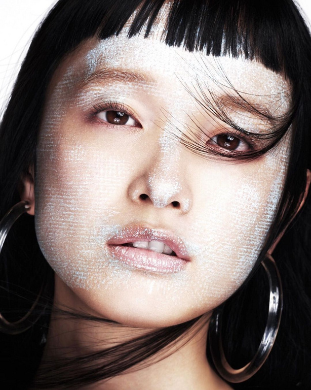 Close-up portrait of an Asian woman with silver makeup, black hair, hoop earrings, and neutral expression against a white backdrop.