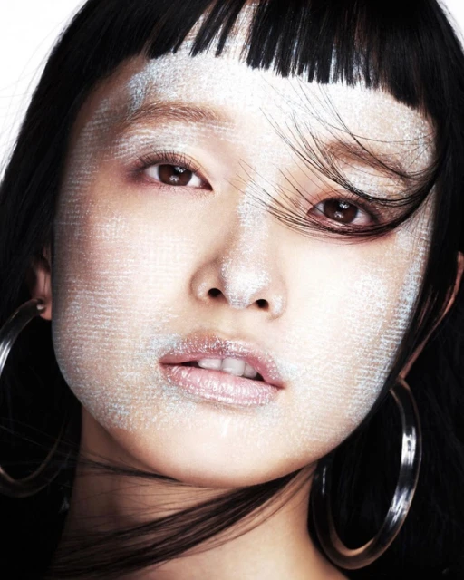 Close-up portrait of an Asian woman with silver makeup, black hair, hoop earrings, and neutral expression against a white backdrop.