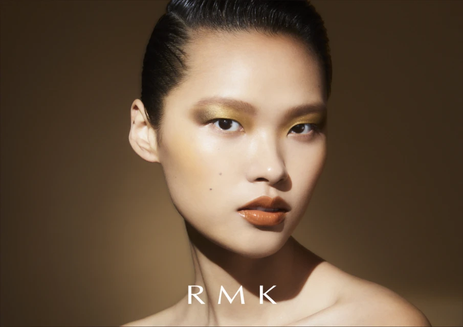 Close-up portrait of an Asian female model with slicked-back hair, golden eye makeup, and glossy lips on a neutral tan background.