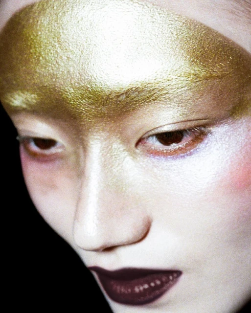Portrait of an adult woman with gold-colored makeup on her forehead and dark-colored lipstick in a beauty studio shot.