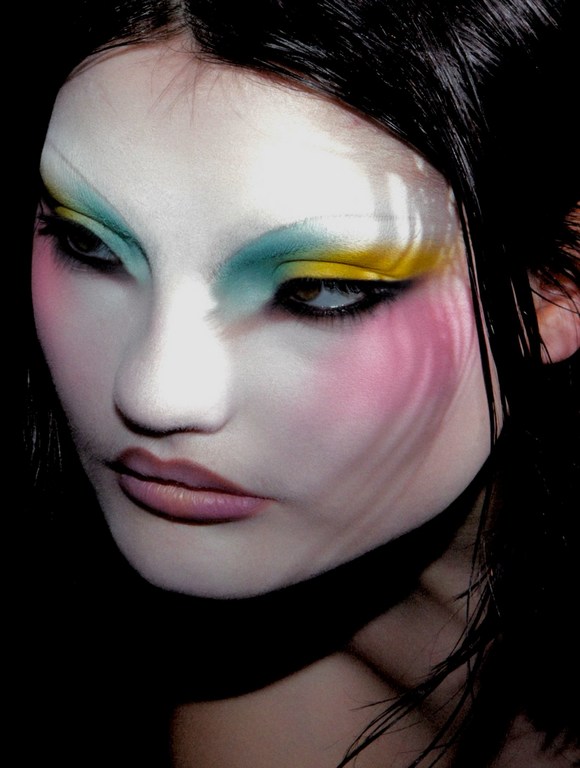 Close-up portrait of a woman with pale skin, wearing colorful makeup in shades of pink, yellow and teal with black hair in a dark setting.