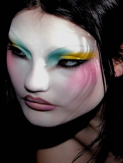 Close-up portrait of a woman with pale skin, wearing colorful makeup in shades of pink, yellow and teal with black hair in a dark setting.