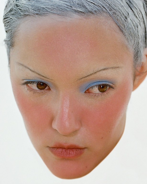 Close-up portrait of an adult female face with light skin, brown eyes, pale pink blush, blue eyeshadow, and light grey hair.