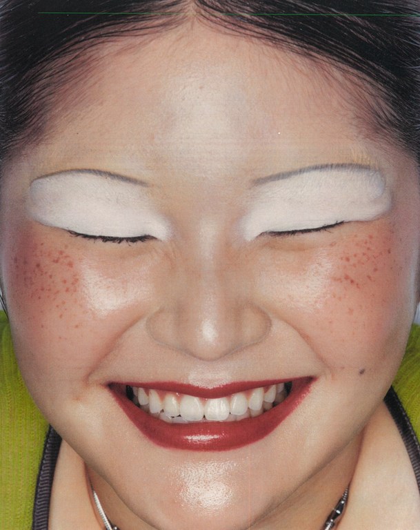 Close-up portrait of a smiling woman with white eyeshadow, red lipstick, and faux freckles by makeup artist Jezz Hill.