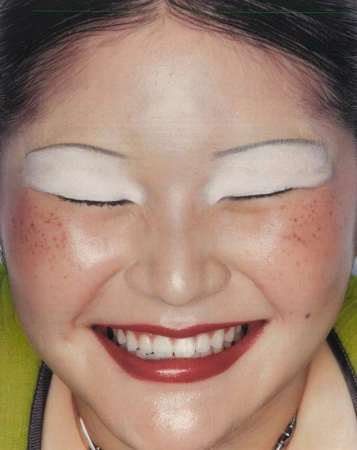 Close-up portrait of a smiling woman with white eyeshadow, red lipstick, and faux freckles by makeup artist Jezz Hill.