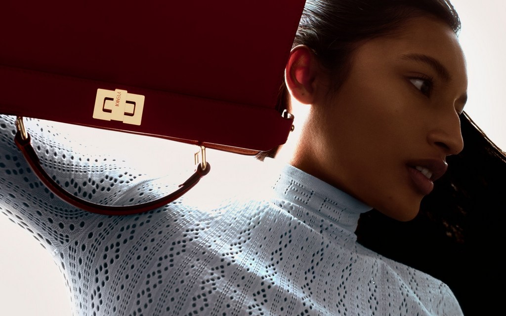 Close-up portrait of a woman wearing a light blue patterned turtleneck holding a red handbag by Fendi against a bright light source
