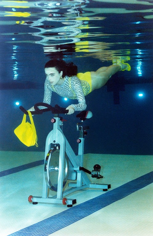 Woman underwater on stationary bike in pool wearing yellow swimsuit and striped long-sleeved shirt, creating a surreal fitness image.