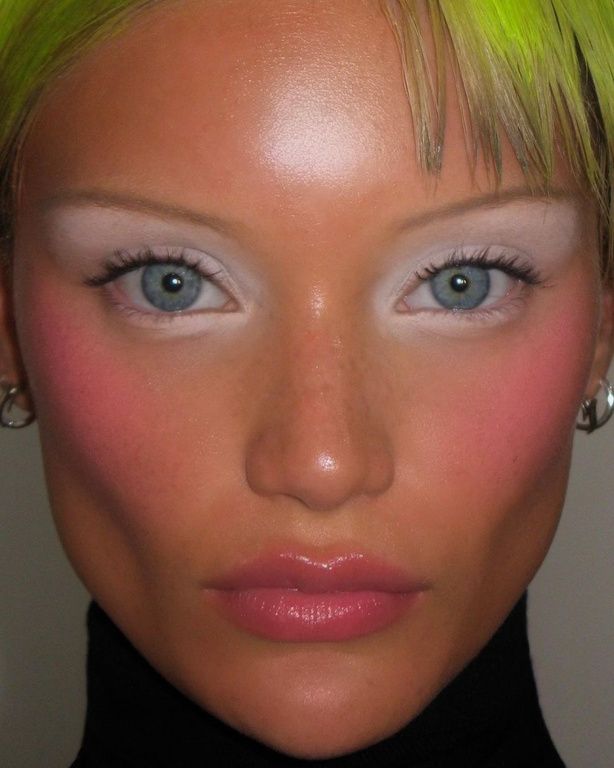 Close-up portrait of a young adult female with striking makeup, including light pink blush, bleached eyebrows, and glossy lips, wearing a black turtleneck.