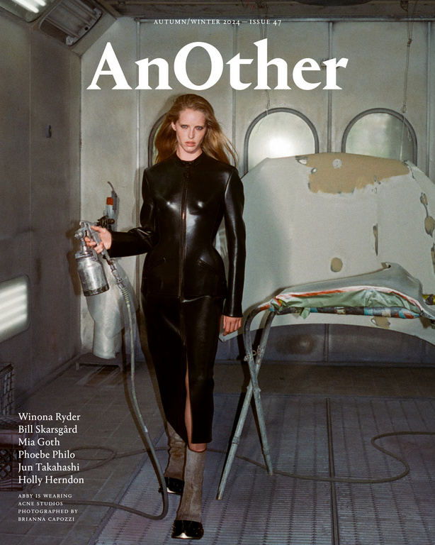Woman in black latex outfit holding spray gun in rundown auto paint booth for high fashion magazine cover shoot with industrial vibe.